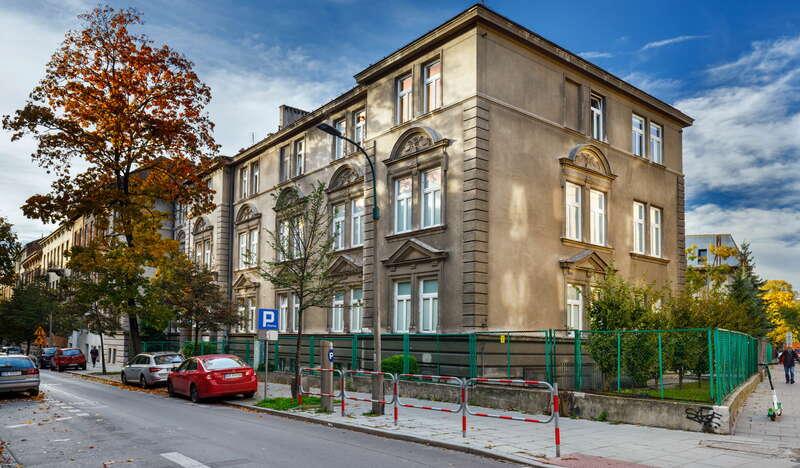 Szary budynek Szpitala Siemiradzkiego w Krakowie. Przy budynku biegnie droga, przy której zaparkowane są samochody. Tuż obok nich widać jesienne drzewo.