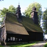 Drewniany ciemny kościół z wysokim, stromym dachem, pokrytym gontem. Na dachu po środku wieżyczka z sygnaturką. Dalej na końcu wysoka wieża z baniastym hełmem. Po lewej stronie przy ścianie schody do wejścia. Na bocznej ścianie powieszone tablice informacyjne i plakaty. Do kościoła prowadzi alejka z kamieni. Wokoło wysokie drzewa. Niebo z lekkimi chmurami.