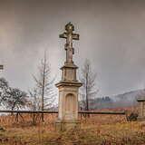 Krzyże greckokatolickie na kamiennych płytach na cmentarzu porośniętym trawą. Z tyłu ogrodzenie z drewnianej belki. Za nim dwa modrzewie bez igieł i obok dwa drzewa. W oddali po prawej zalesione wzgórze. Szare niebo i opadająca mgła.