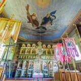 Widok na wnętrze cerkwi od dołu. Po prawej i lewej drewniane ławki z chorągwiami. Na wprost bogato zdobiony ikonostas, częściowo zasłonięty u góry kryształowym żyrandolem. Na suficie malowany duży obraz Trójcy świętej.