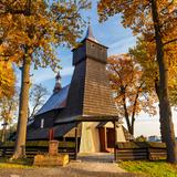 Drewniany kościół z wysoką wieżą otoczony drewnianym ogrodzeniem oraz kilkoma drzewami w pomarańczowo-żółtych barwach jesieni. Widok od strony wejścia do świątyni.