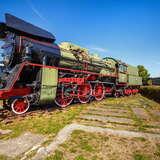 Pogodny dzień. Piękna czarno-zielona zabytkowa lokomotywa z czerwonymi kołami w Skansenie Taboru Kolejowego w Chabówce. Przed nią betonowe płyty porośnięte trawą z niskim żywopłotem. Po bokach widać wagony.