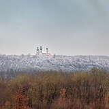 Widok z oddali na wieże klasztorne. Z przodu las w zimowej szacie, nagie drzewa, z tyłu zaśnieżone wzgórze.