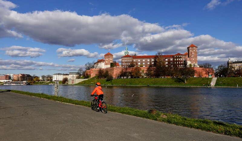 Widok na osobę jadącą na rowerze wzdłuż bulwarów w Krakowie. W tle widok na zamek Królewski na Wawelu.