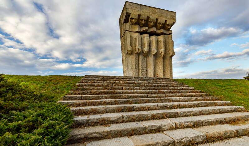 Betonowy pomnik z sylwetkami pięciu postaci na obszarze byłego obozu KL Płaszów w Krakowie. Prowadzą do niego kamienne schody, obok nich trawa, a nad tym niebieskie, zachmurzone niebo.